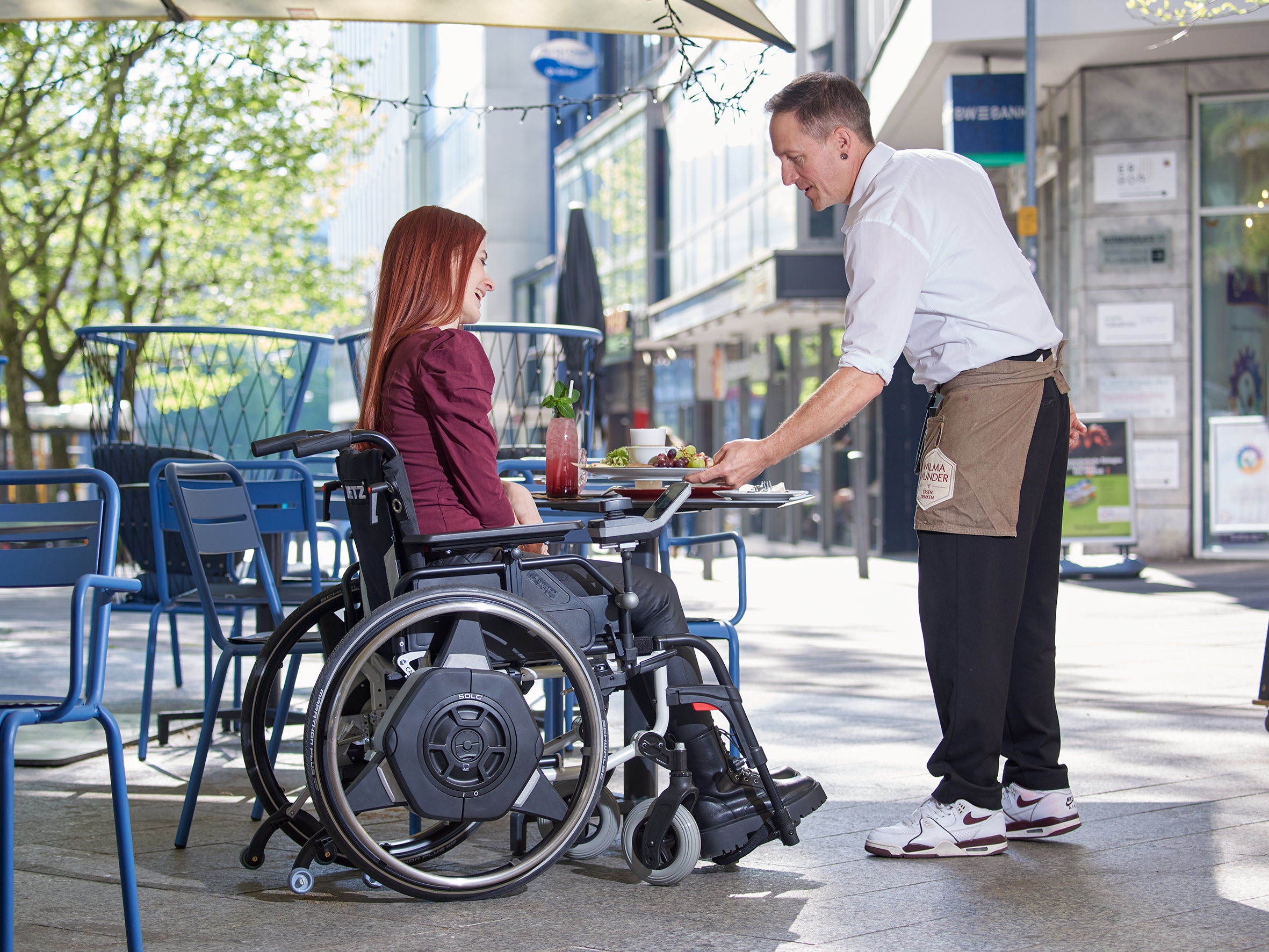 Das Bild zeigt eine junge Frau, die mit ihrem manuellen Rollstuhl und Zusatzantrieb für Rollstühle SOLO² auf einer Terrasse eines Cafès in Stuttgart (Deutschland) sitzt. Der Kellner stellt ihr gerade einen Teller mit Frühstück auf ihren Tisch. Zu sehen ist außerdem die Seitenansicht des Rollstuhlantriebs SOLO² der Firma AAT sowie das Bediengerät für die Steuerung.